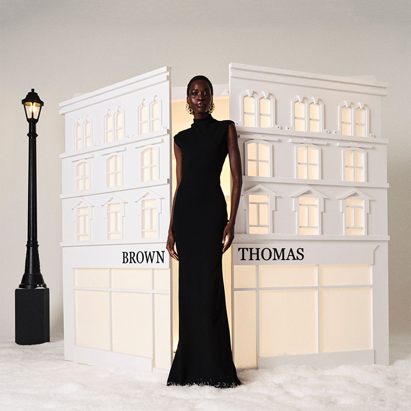 A person in a long black dress stands in front of a model building labeled 'Brown Thomas' beside a streetlamp.

