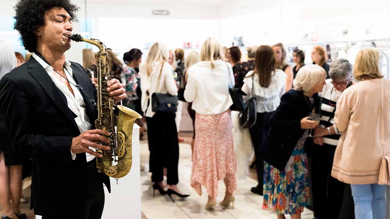 A saxophonist in a suit plays music at a social event with a crowd of people in a well-lit indoor setting.